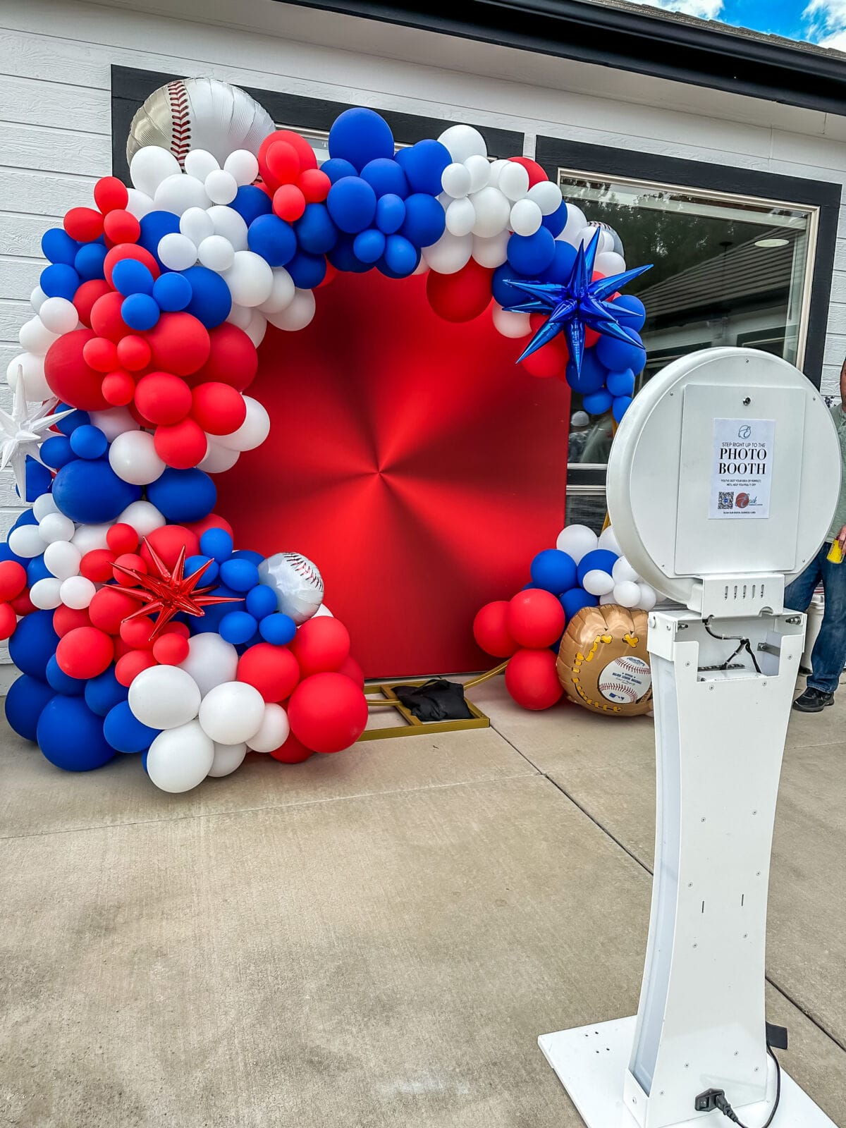 Red, white, and blue baseball balloon arch beside a photo booth stand outdoors