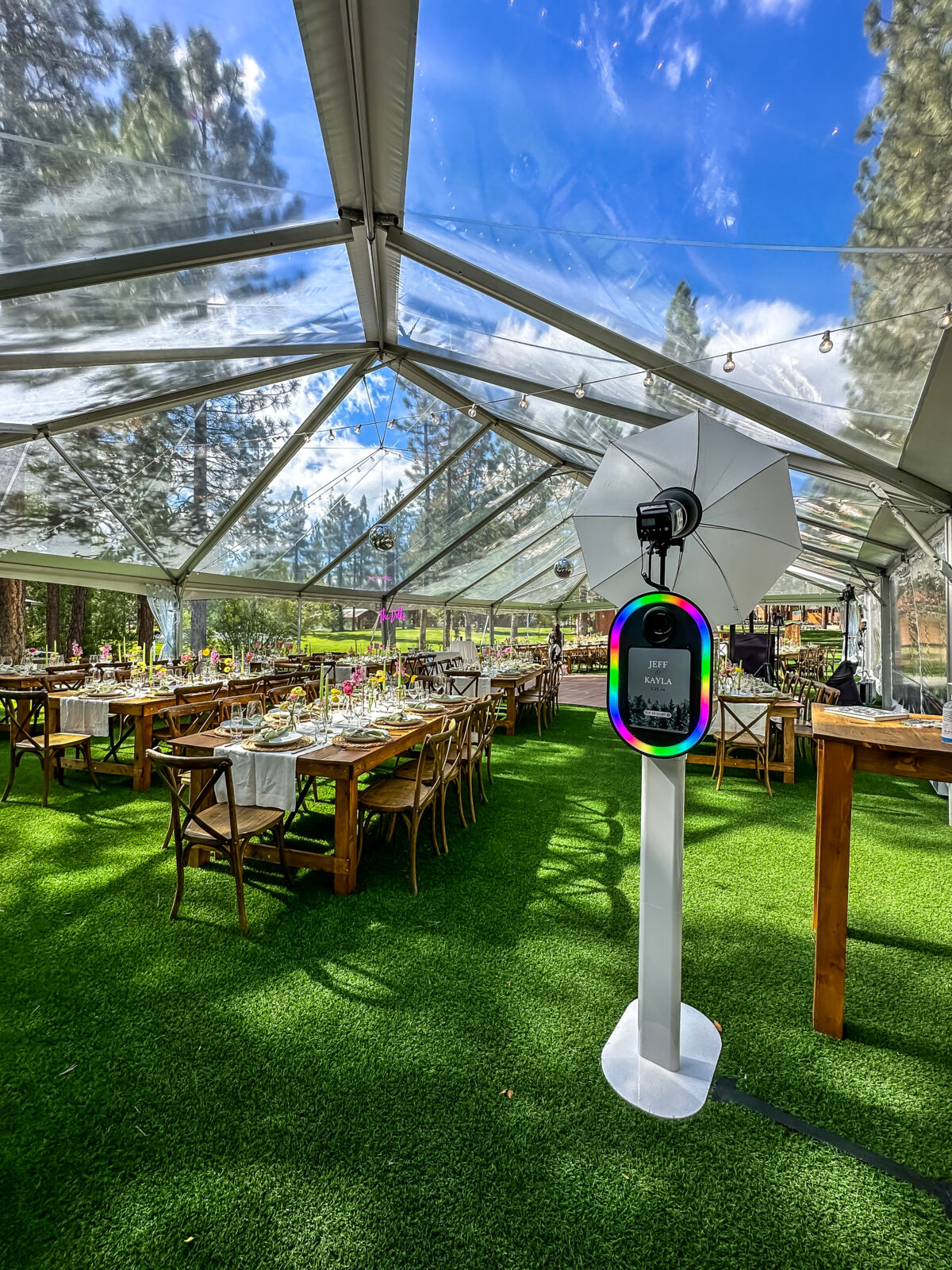 Photo booth set up inside a clear-top tent reception on green grass with Tahoe pines overhead