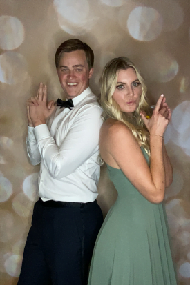 Two guests posing back-to-back with finger-gun poses against a gold bokeh photo booth backdrop