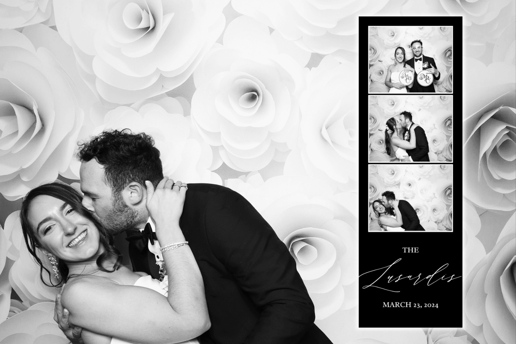 Black and white photo of a couple kissing at a white paper-flower wall