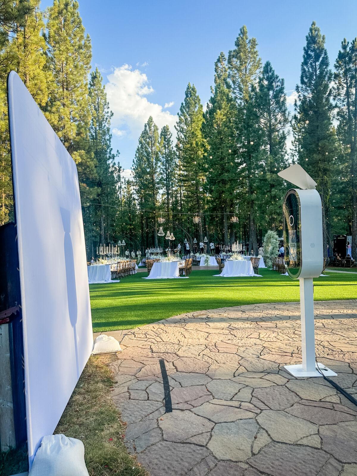 Outdoor photo booth with a white backdrop on a stone patio surrounded by pine trees