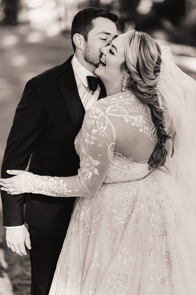 Black and white photo of a groom kissing a laughing bride’s cheek, her beaded gown and veil flowing