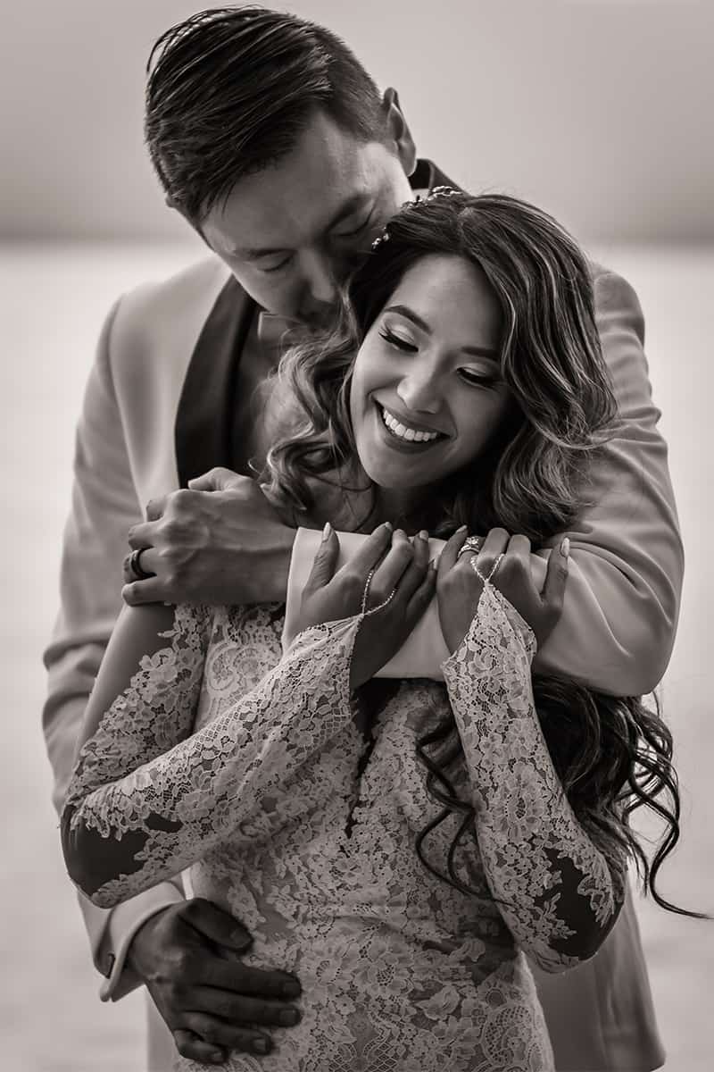 Black and white portrait of a groom embracing a smiling bride from behind