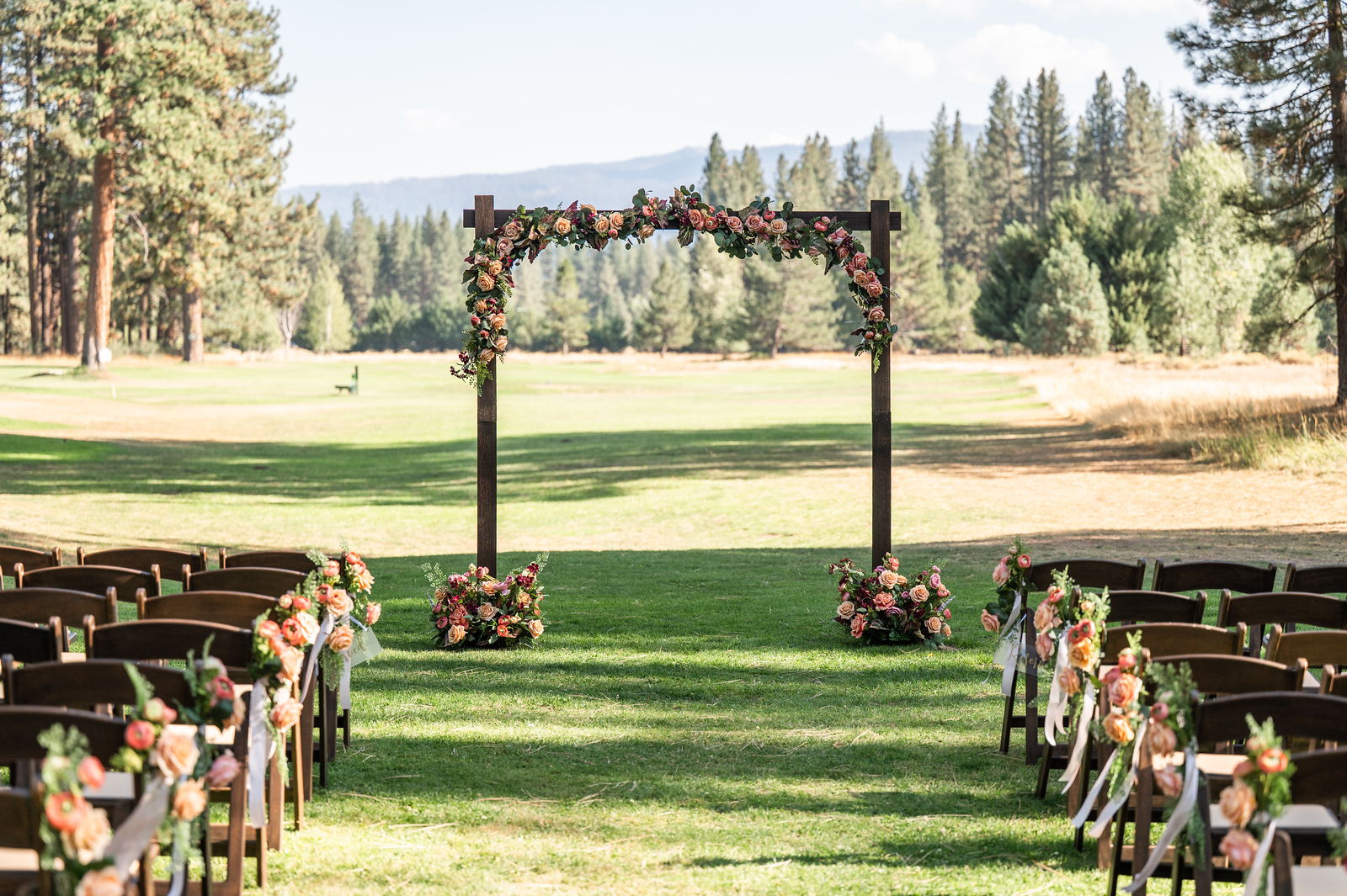 Empty ceremony setup with a wooden floral arch on a golf-course lawn framed by pines and mountains