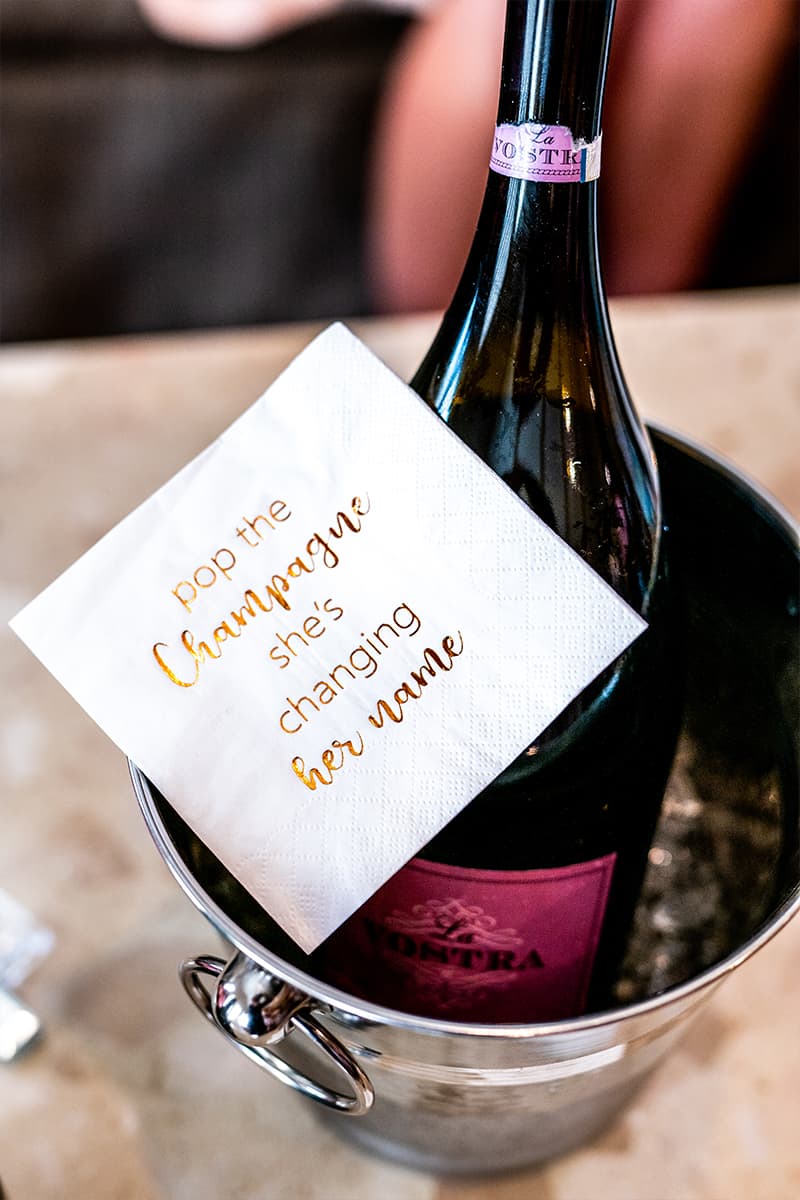 Champagne bottle in an ice bucket with a gold-foil pop the champagne she’s changing her name napkin