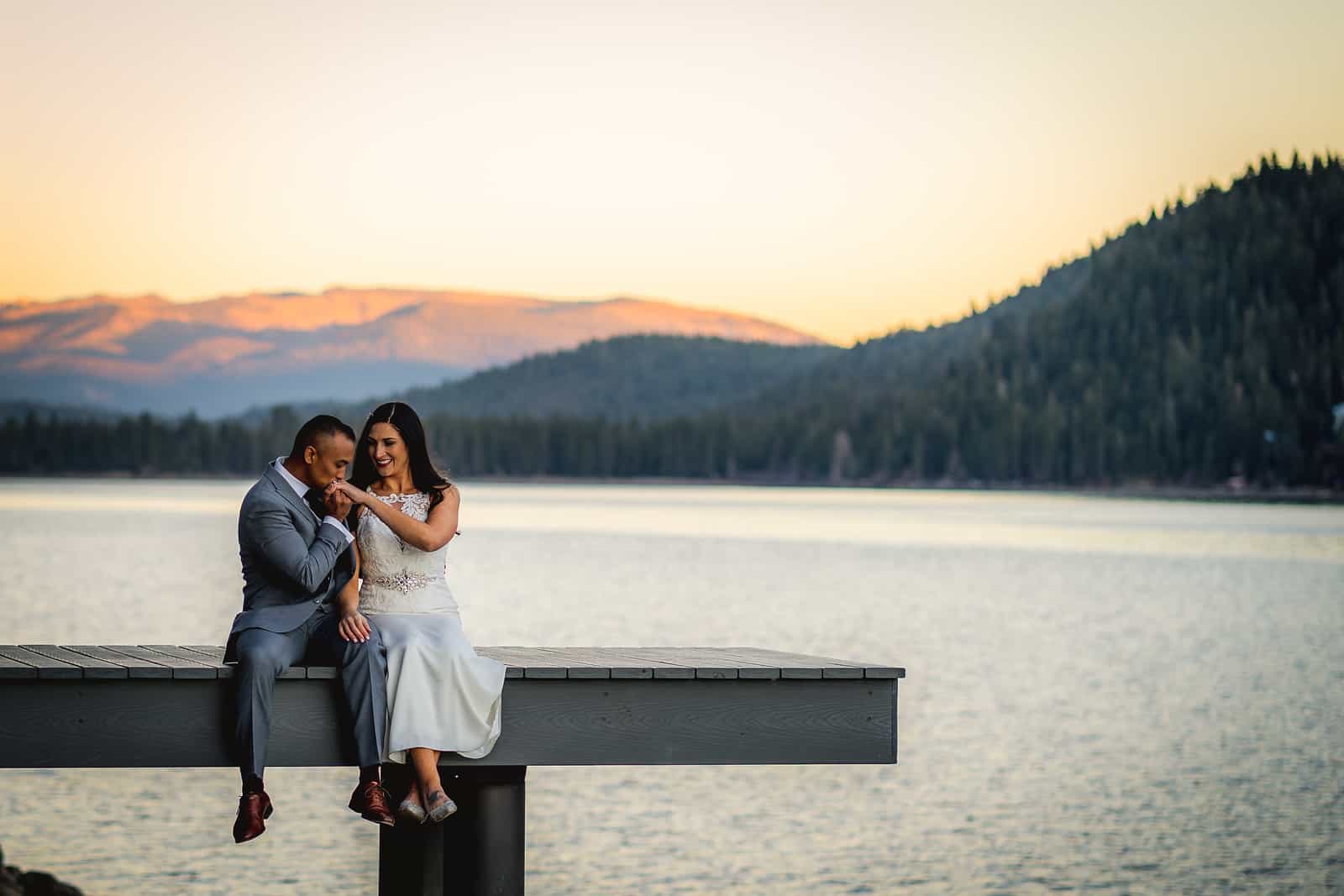 Groom kissing the bride’s hand while seated on a lakeside dock at dusk with mountains behind