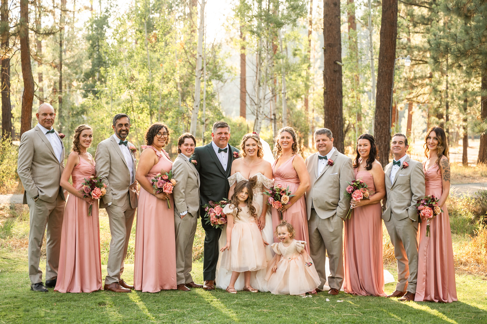 Full wedding party in pink and tan attire among tall pines with two flower girls