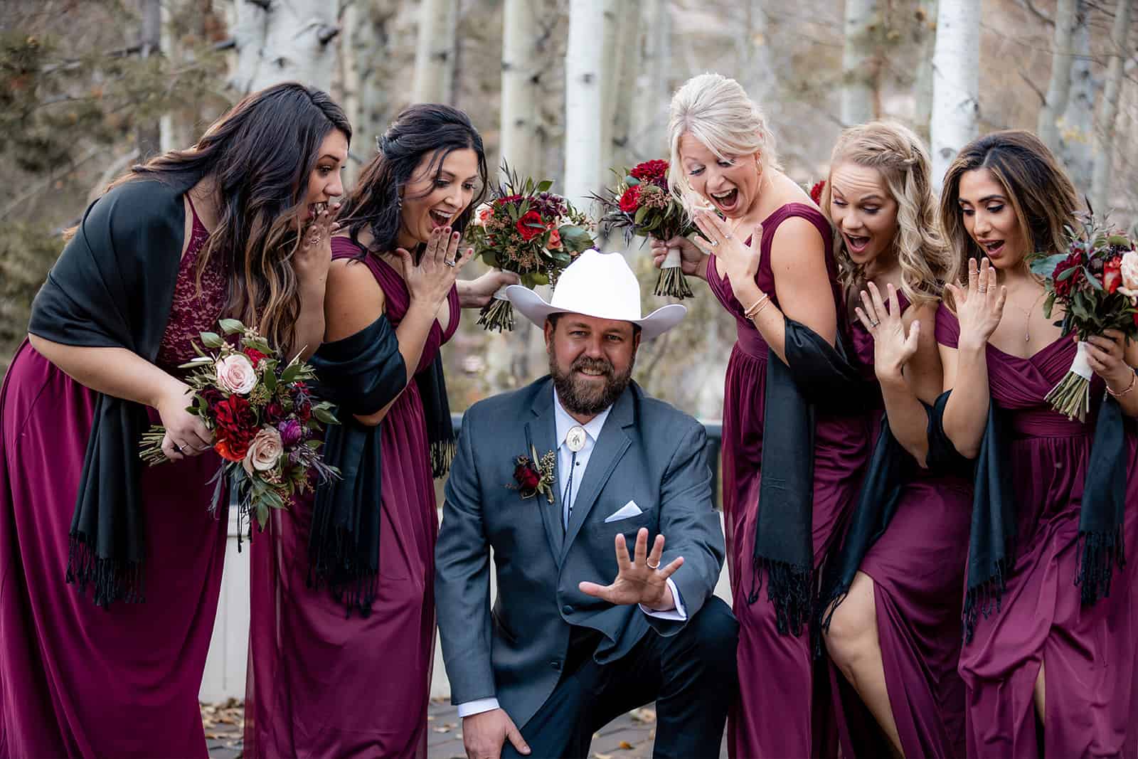 Groom in a white cowboy hat kneeling while burgundy-clad bridesmaids react and laugh