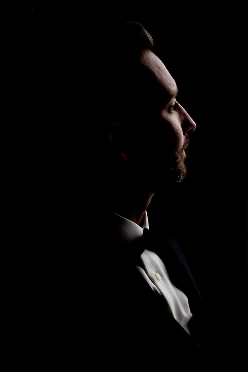 Dramatic low-key profile portrait of the groom in a tuxedo against a black background