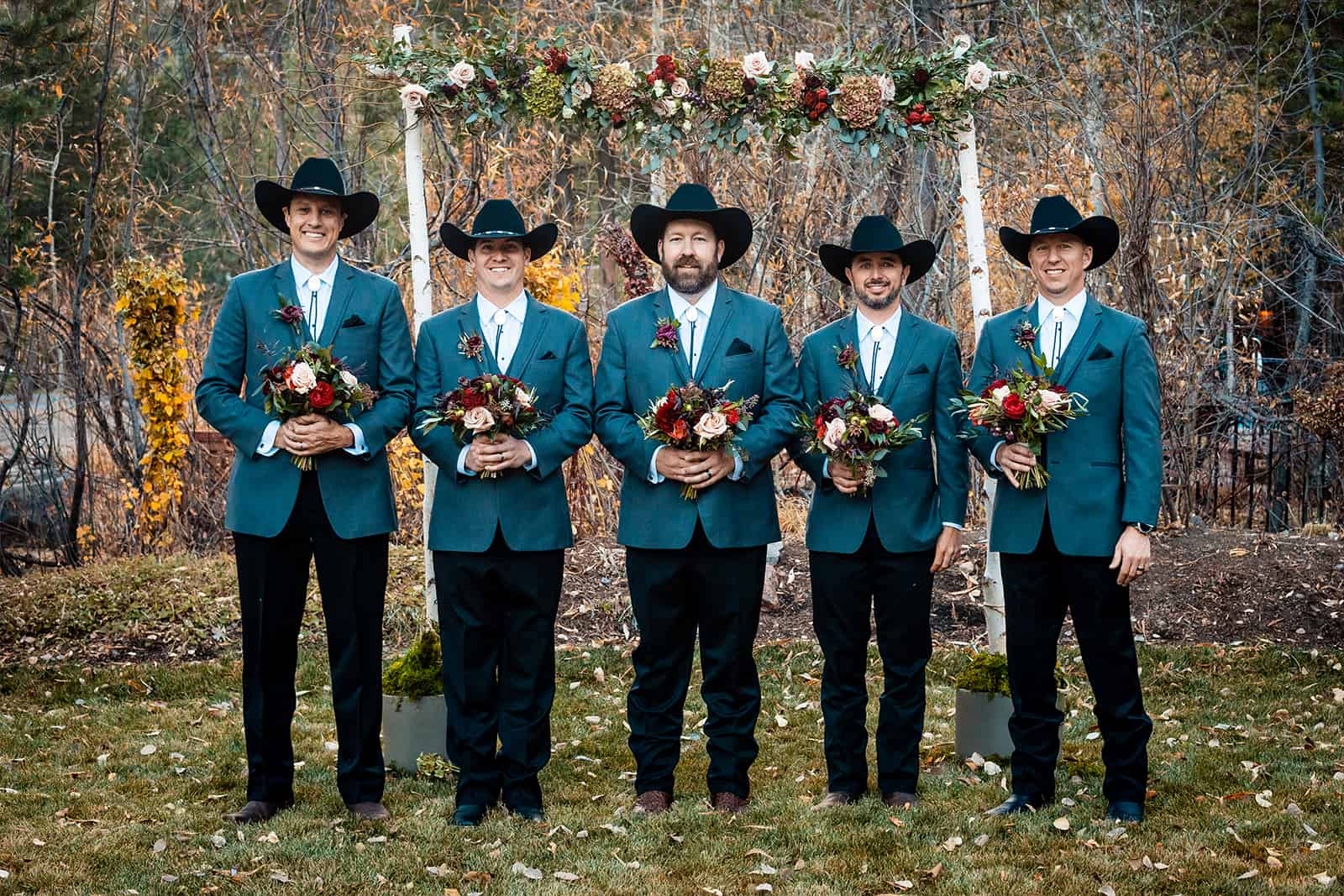 Five groomsmen in teal suits and black cowboy hats holding bouquets under a fall floral arch