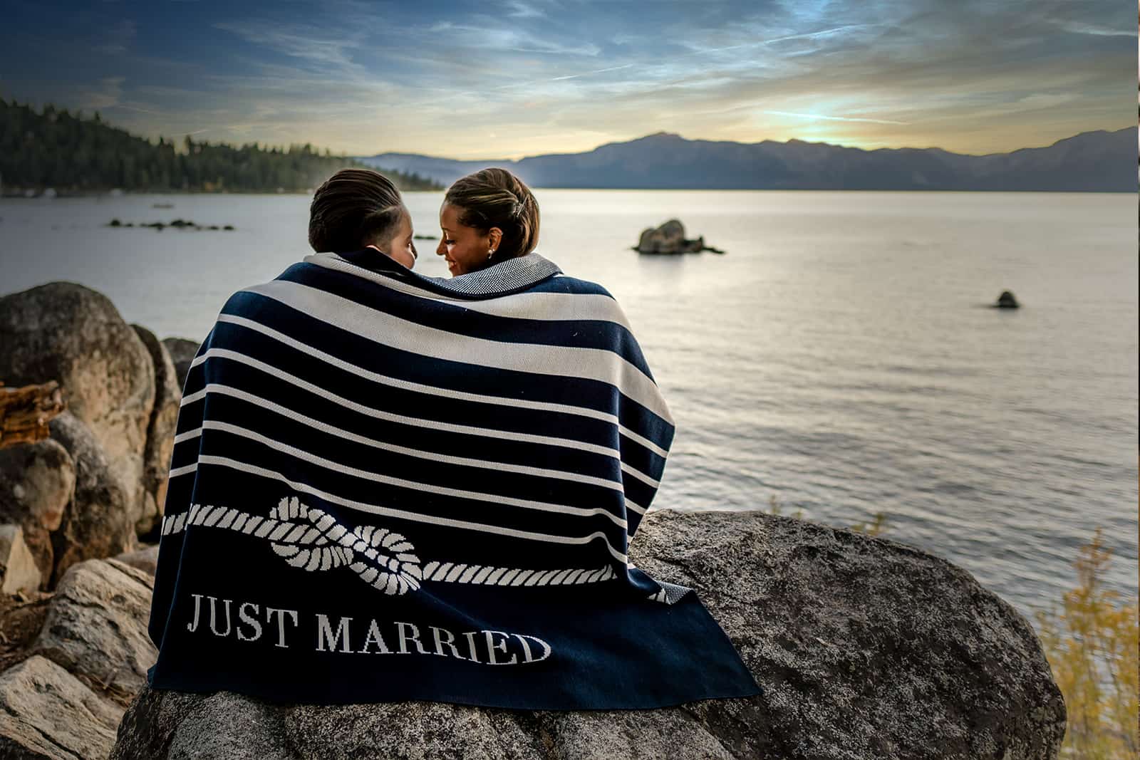 Couple wrapped in a navy Just Married blanket on a rock overlooking Lake Tahoe at dusk