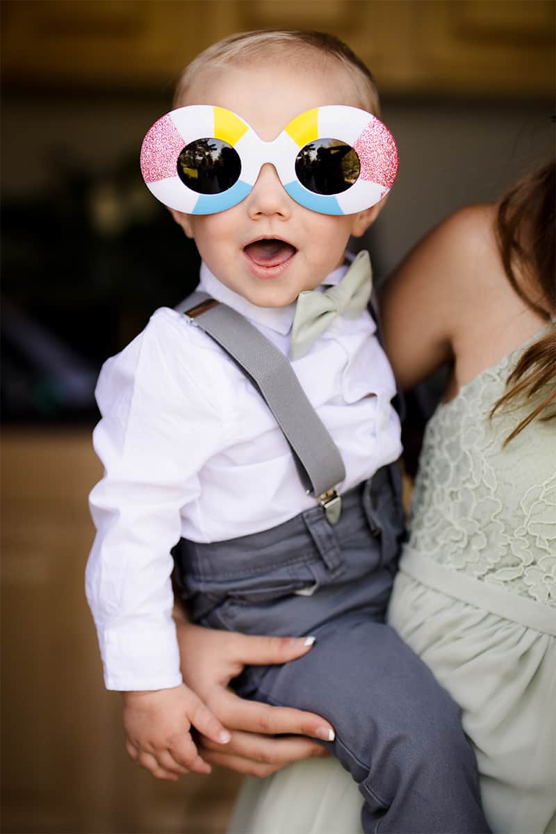 Ring bearer toddler in suspenders and a sage bow tie wearing oversized colorful novelty sunglasses